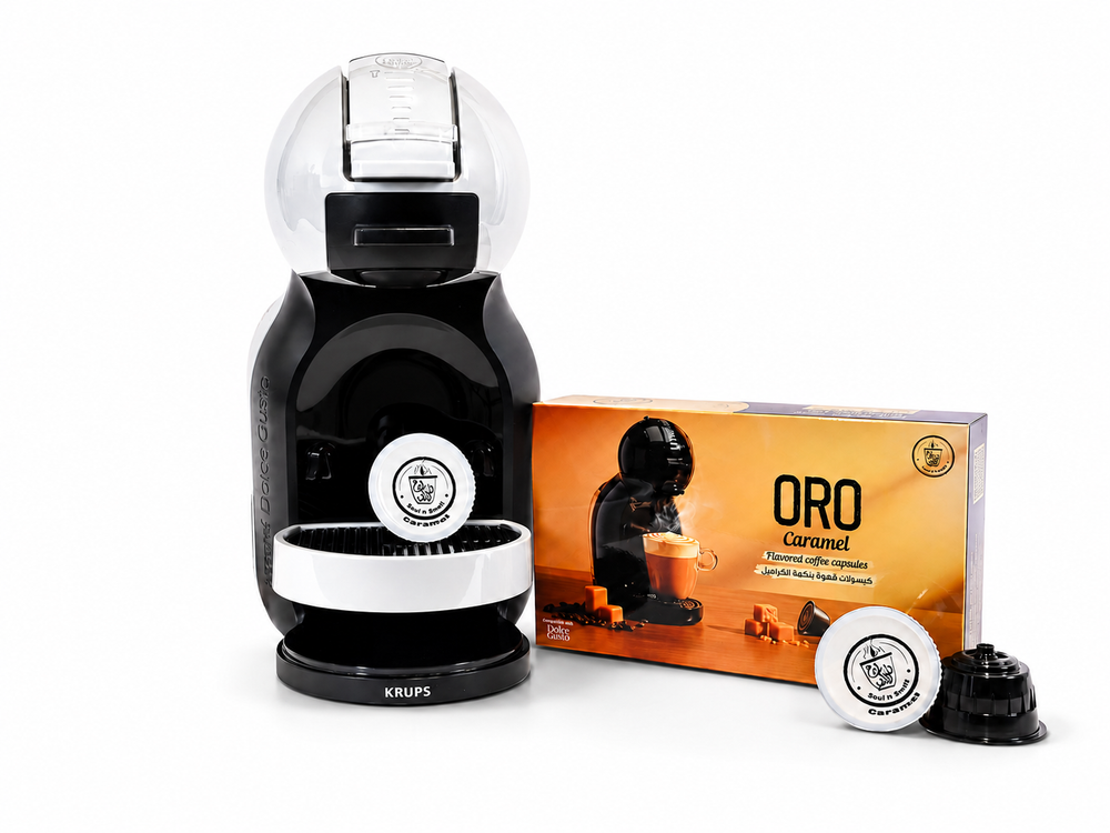 ORO - Caramel Cappuccino - A black and white Krups Nespresso capsule coffee machine shown next to its orange ORO Caramel coffee capsule packaging box, with two coffee capsules (one filled with coffee and one empty) in front, on a white background.